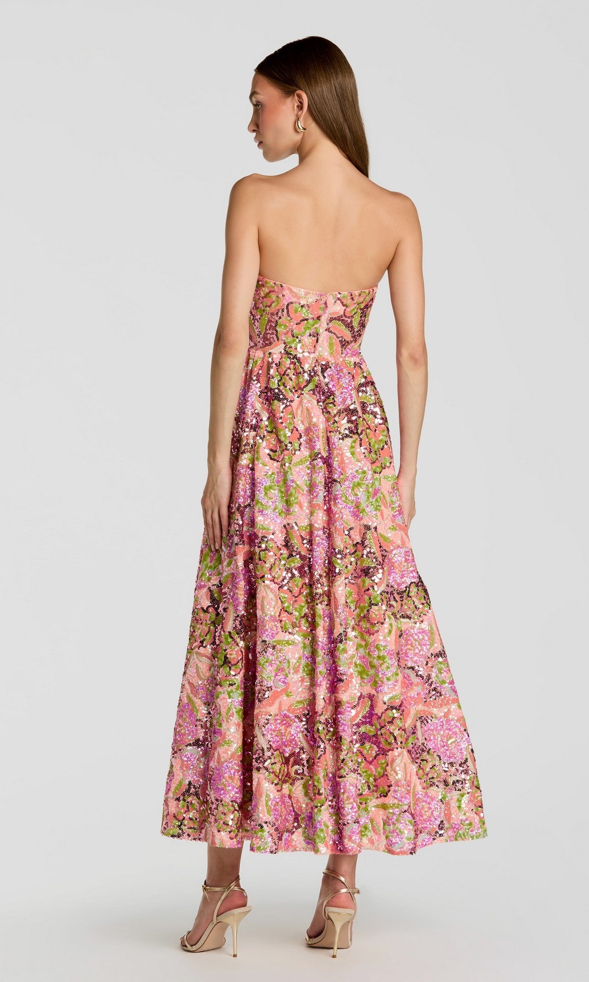 A woman stands facing away from the camera against a plain light background, wearing the Tea-Length Party Dress: Mac Duggal 56386—a strapless, pink floral sequin party dress paired with high heels.